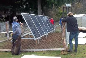Grupo de personas observando la instalación de paneles solares montados sobre una estructura metálica dentro de un área cercada. Al fondo, árboles y una pequeña construcción sugieren un entorno rural o semirrural.